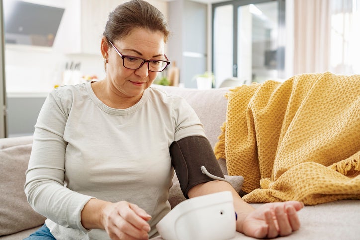 Mature woman sitting on sofa using digital blood pressure gauge to measure high blood pressure.